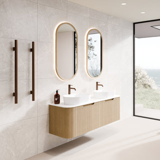 Modern floating double-sink bathroom vanity with two timber oval backlit mirrors, bronze wall-mounted handles, a freestanding bathtub, and large windows overlooking the ocean.