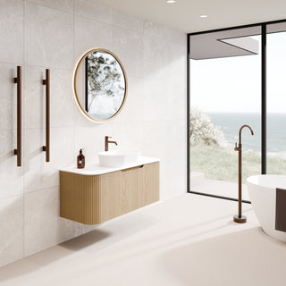 Modern floating bathroom vanity with a white rectangular sink, ribbed wood cabinet, round timber-framed mirror, and matte bronze wall-mounted fixtures in a bright coastal bathroom.
